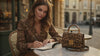 A model representing LU DI LUSSO enjoying a creative moment at a European café, with the iconic handbag next to her.